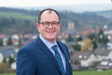 Bild von Bürgermeister Marian Knauf in blauem Anzug und weißem Hemd und blauem Schlips in der Mitte des Bildes. Der Hintergrund ist unscharf und man kann Neukirchen erahnen.