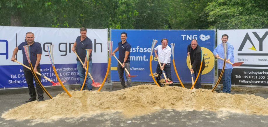 Foto (v. l. n. r.): Bekir Turan (Geschäftsführer TBG GmbH), Raphael Kupfermann (TNG-Regionalleiter), Juan Torp (TNG-Netzplaner), Sükrü Tasdemir (Projektleiter Aytac Bau GmbH), Marian Knauff (Bürgermeister Neukirchen), Jörn Szypkowski (Digitalisierungsbeauftragter Neukirchen). stehen mit Spaten hinter einen Haufen Sand. Im Sand stecken kurze Stücke von Glasfaserleerrohren.