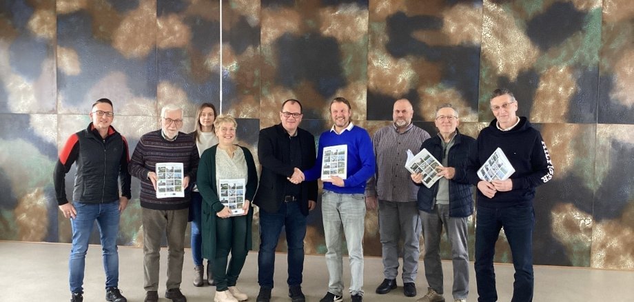 Die IKEK Steuerungsgruppe von links Thorsten Brill (Steuerungsgruppe), Helmut Reich (Steuerungsgruppe), Ljuba Iljin (Sachbearbeiterin), Nicole Mattheis (Steuerungsgruppe), Bürgermeister Marian Knauff, Marco Link (Arbeitsgruppe Stadt), Sven Hübl (Steuerungsgruppe), Willi Berg (Steuerungsgruppe), Hartwig Hepp (Steuerungsgruppe) steht im Stadtverordnetensitzungssaal der Stadt Neukirchen. Einige Steuerungsmitglieder halten das Stadtentwicklungskonzept in der Hand.